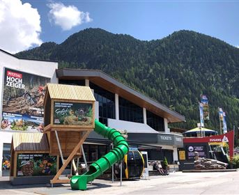 A modern ticket building with a large play tower and a long slide. In the background, green mountains and a clear sky are visible.