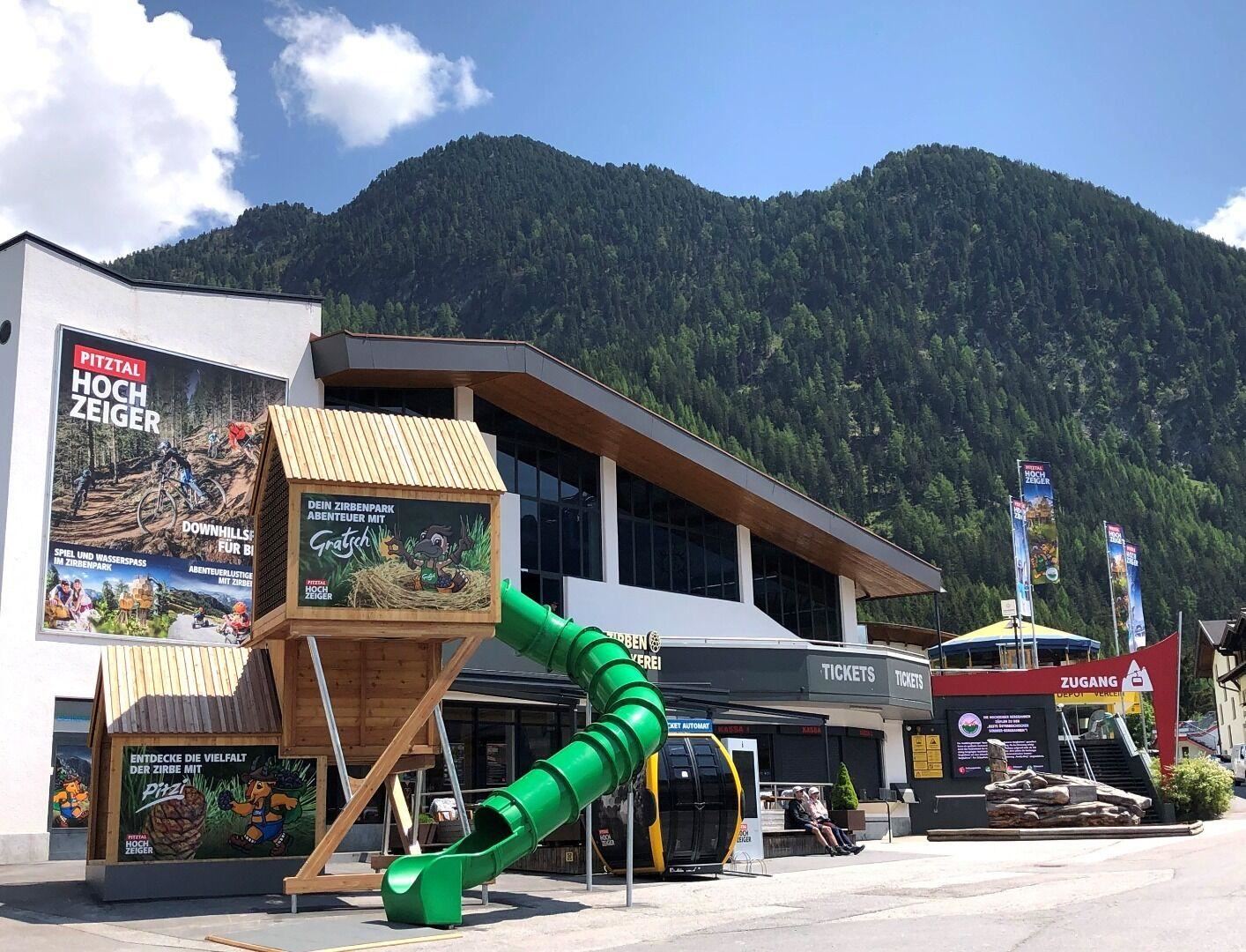 A modern ticket building with a large play tower and a long slide. In the background, green mountains and a clear sky are visible.