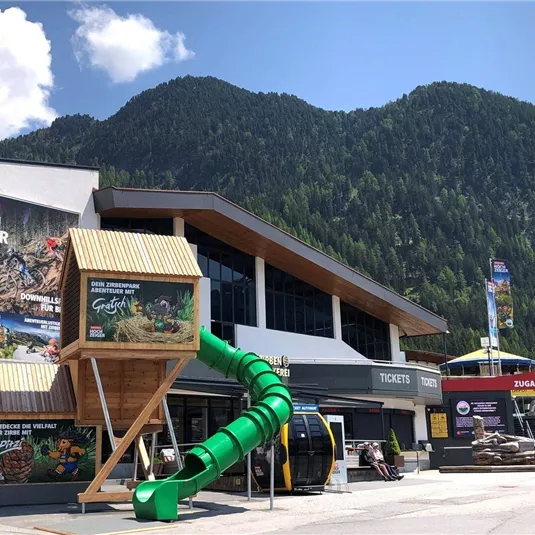 A modern ticket building with a large play tower and a long slide. In the background, green mountains and a clear sky are visible.