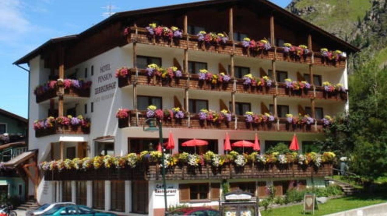 Ein charmantes Hotel mit sichtbaren Balkonen, die mit bunten Blumen geschmückt sind. Der Himmel ist klar und die Umgebung ist grün.