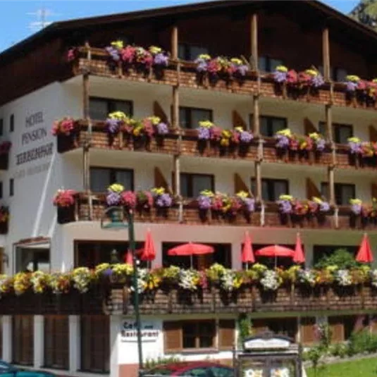 Ein charmantes Hotel mit sichtbaren Balkonen, die mit bunten Blumen geschmückt sind. Der Himmel ist klar und die Umgebung ist grün.