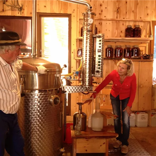 Eine Person erklärt die Destillation in einer Holzwerkstatt. Im Hintergrund sind Flaschen und Geräte zur Herstellung sichtbar.