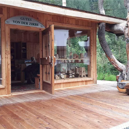 Ein Holzladen mit großen Fenstern und einer einladenden Tür. Draußen steht ein Wagen und im Hintergrund sind Bäume zu sehen.