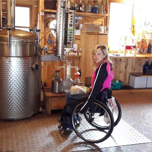 Eine Person im Rollstuhl sitzt in einem gemütlichen Raum mit einer großen, silbernen Destillationsapparatur. Der Raum ist rustikal eingerichtet, mit Holz und verschiedenen Utensilien.