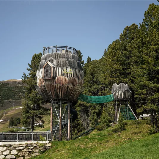 Zwei Baumhäuser, die wie große Blumen gestaltet sind, stehen auf einem Hügel umgeben von grünen Bäumen. Im Hintergrund sind Berge und ein klarer blauer Himmel zu sehen.