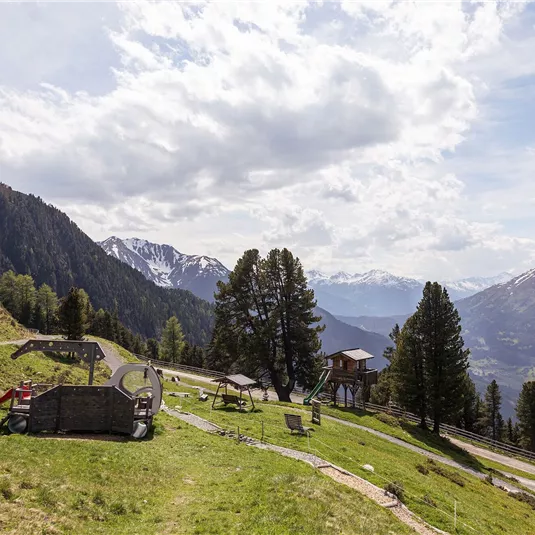 Eine malerische Berglandschaft mit grünen Wiesen und einem Spielplatz. Im Hintergrund sind hohe Berge und ein blauer Himmel mit wenigen Wolken zu sehen.
