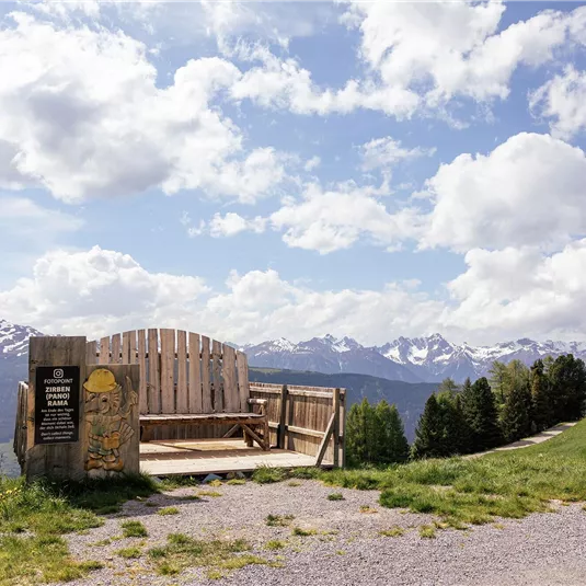 Eine wunderschöne Aussichtsbank auf einem Berg mit Blick auf die schneebedeckten Gipfel. Der Himmel ist klar und von Wolken durchzogen.