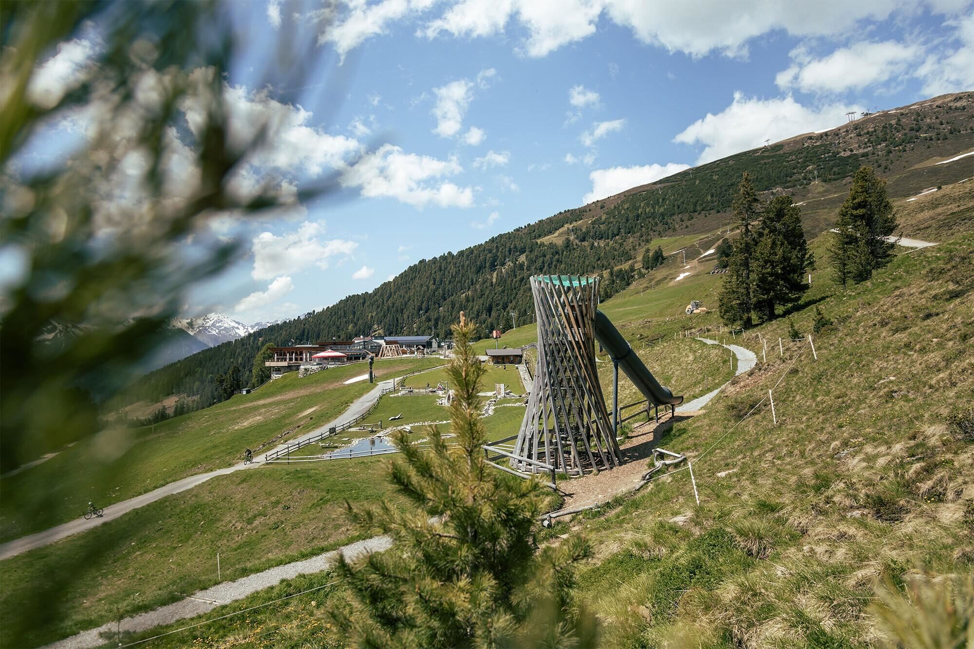 Ein schöner Spielplatz in den Bergen mit einer Rutsche und einer beeindruckenden Holzstruktur. Umgeben von grünen Wiesen und einem klaren Himmel.