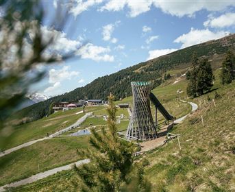 Eine grüne Alm mit einem Spielplatz in der Mitte. Im Hintergrund sind Berge und ein blauer Himmel zu sehen.