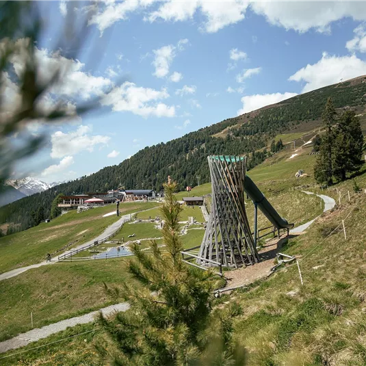 Eine grüne Alm mit einem Spielplatz in der Mitte. Im Hintergrund sind Berge und ein blauer Himmel zu sehen.