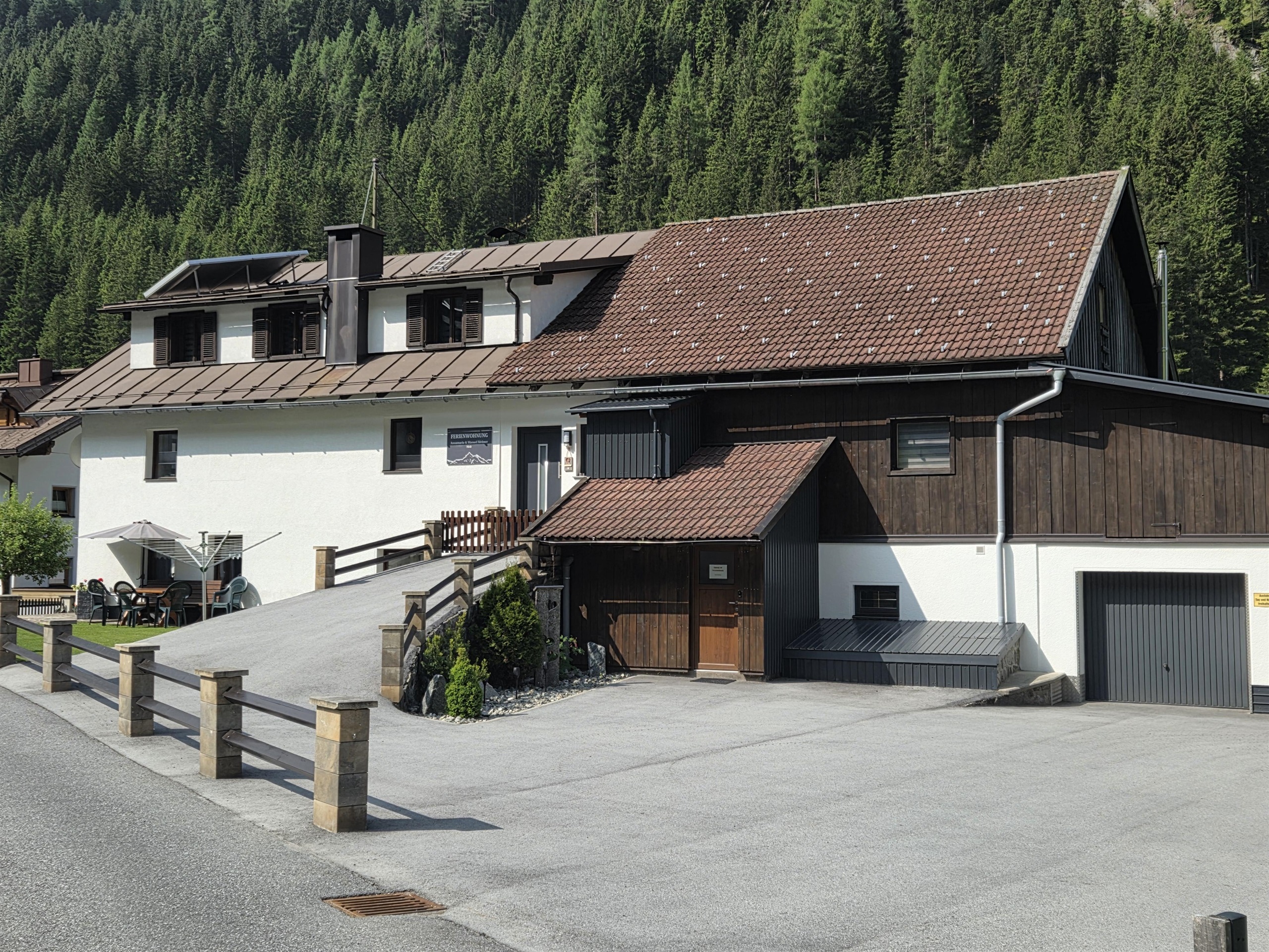 Ein großes, zweistöckiges Haus mit einem geneigten Dach und einer Mischung aus Holz- und Putzfassade. Im Hintergrund sind grüne Berge zu sehen und es gibt eine asphaltierte Zufahrt vor dem Gebäude.