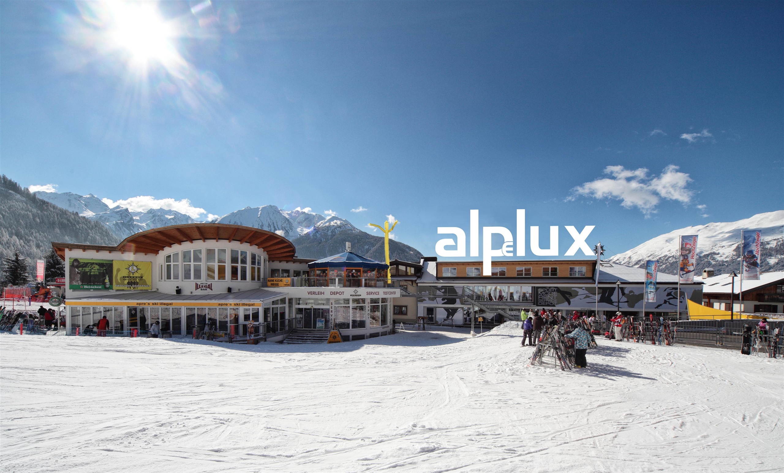 A sunny day on the slopes in the mountains. In the foreground, a modern building with the inscription "alplux" and snow on the ground.