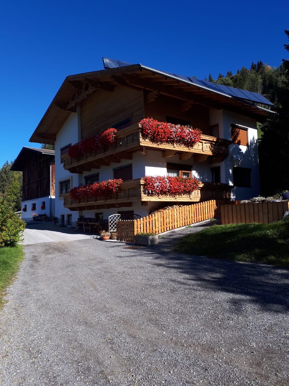 Ein schönes, zweigeschossiges Haus mit Balkonen, die mit roten Blumen dekoriert sind. Es liegt umgeben von Bäumen unter einem klaren blauen Himmel.