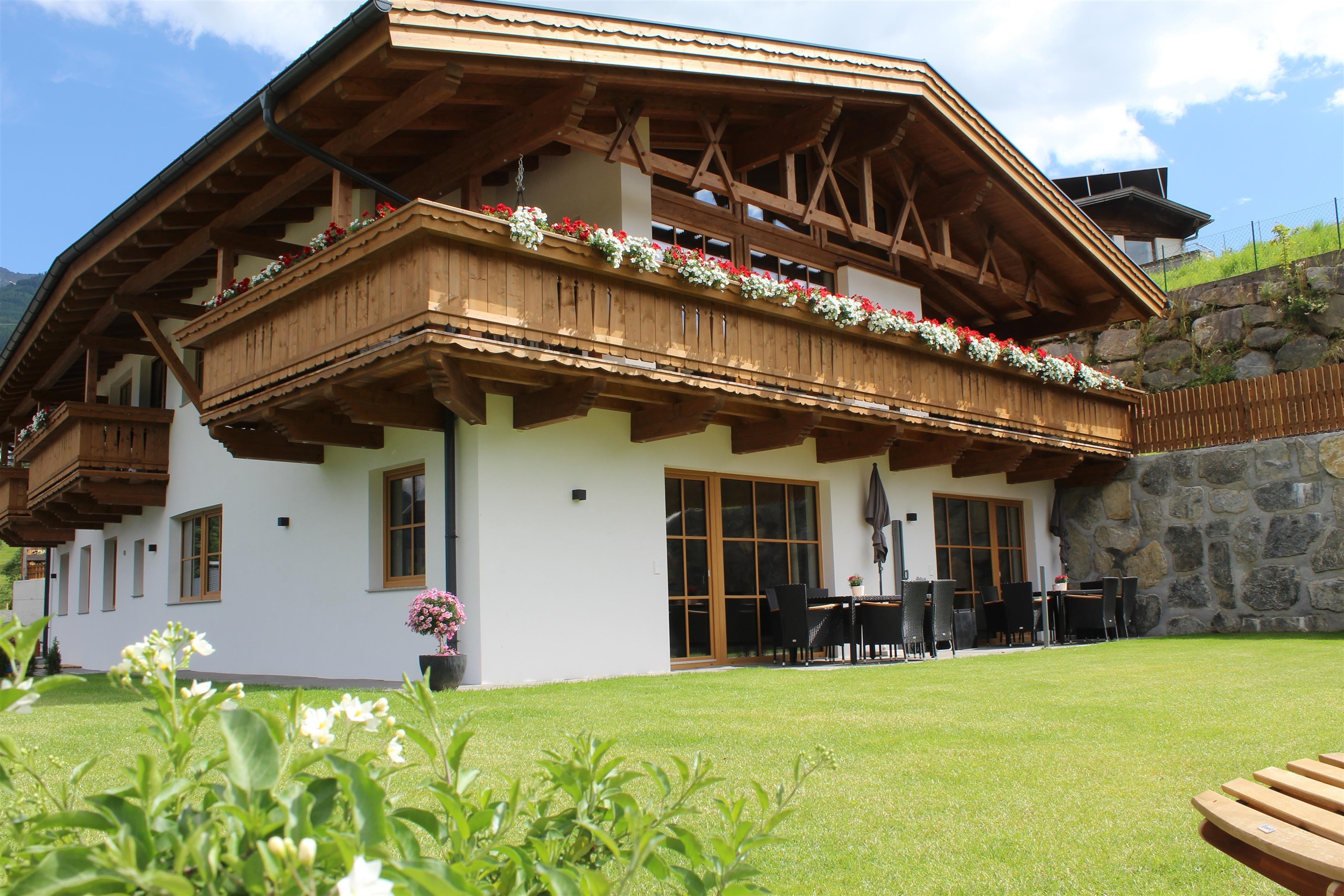 Ein traditionelles alpenländisches Haus mit einem großen Balkon und vielen Blumen. Der Garten ist grün und großzügig angelegt, ideal zum Entspannen.