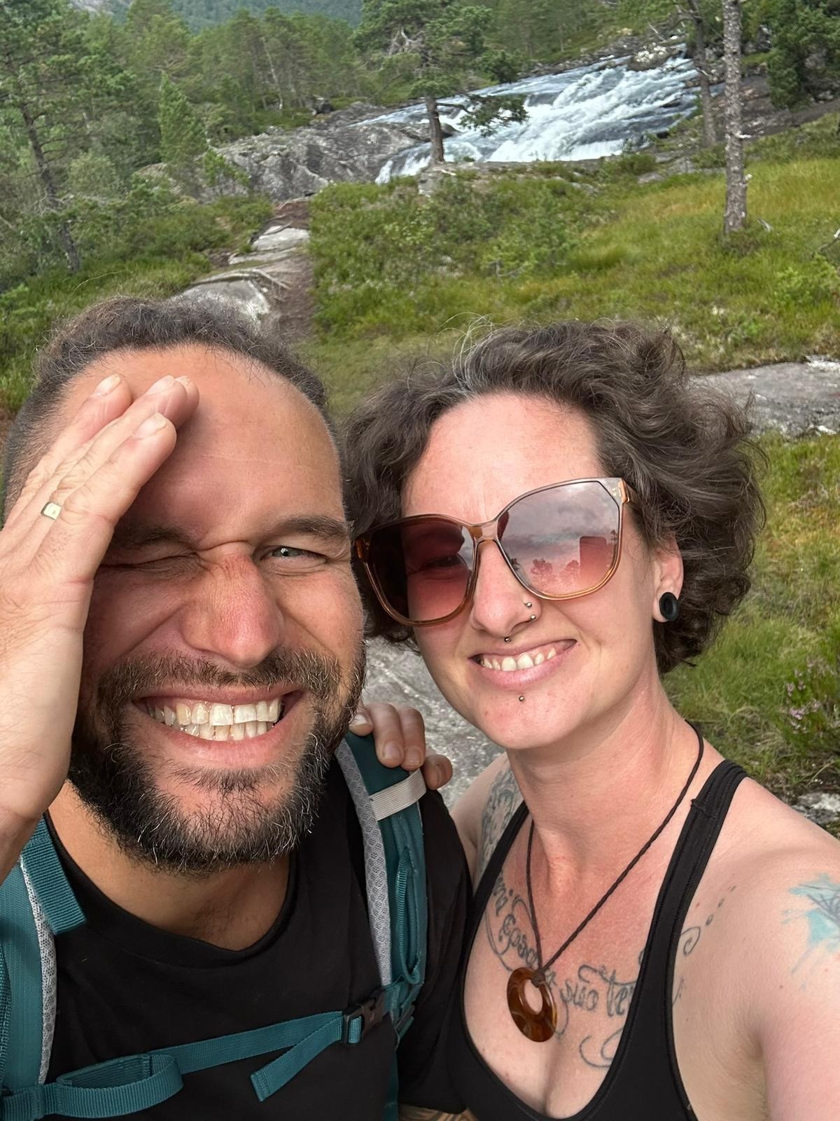 Ein Paar lächelt in der Natur und posiert für ein Foto. Im Hintergrund sind grüne Landschaften und ein Wasserfall zu sehen.