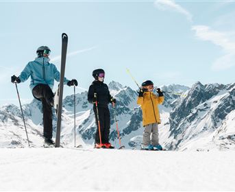 Three skiers are standing on a snow-covered slope in the mountains. The surroundings are bright and sunny, with impressive mountain peaks in the background.