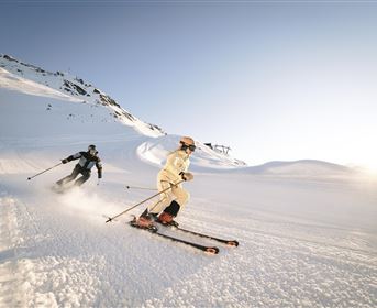 Two skiers glide over a snow-covered slope. In the background, there are mountains and a clear sky.