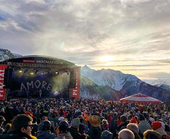 A large audience gathers in front of a stage in the mountains. In the background, snow-capped mountains and a beautiful sky can be seen.