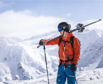 A skier stands on a snow-covered mountain peak and looks into the distance. The surroundings consist of majestic, snow-covered mountains and a clear sky.
