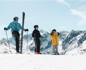 Drei Skifahrer stehen auf einem schneebedeckten Hang und genießen die Aussicht auf die Berge. Die Sonne scheint und der Himmel ist klar.