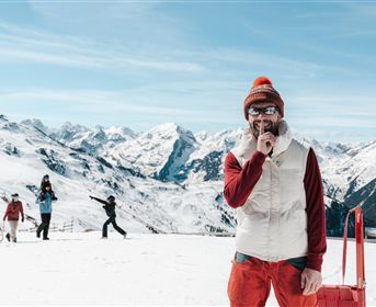 A man in warm clothes stands in the snow and makes a "shhh" sign. In the background, people are playing in the winter area, and snow-covered mountains can be seen.