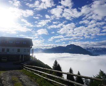 A house on a hill above a blanket of clouds overlooking the mountains. The sky is clear and blue with some white clouds.