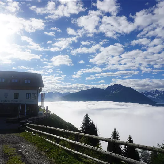 Ein Haus auf einem Hügel über einer Wolkendecke mit Blick auf die Berge. Der Himmel ist klar und blau mit einigen weißen Wolken.