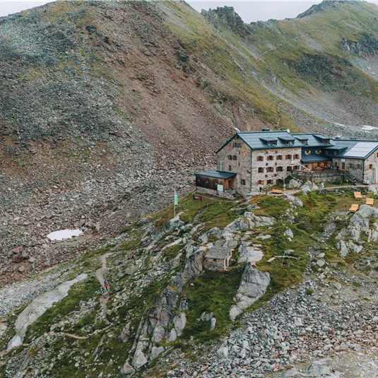 A mountain house on rocky terrain, surrounded by impressive mountains. The landscape is rugged and untamed, with few patches of snow.