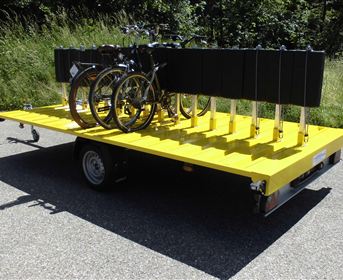 Ein gelbes Anhänger mit mehreren Fahrrädern auf der Rückseite steht auf einer asphaltierte Fläche. Im Hintergrund sind grüne Sträucher und Bäume zu sehen.