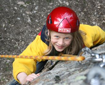 Ein junges Mädchen klettert an einem Felsen und trägt einen Helm. Sie lächelt und scheint viel Spaß zu haben.