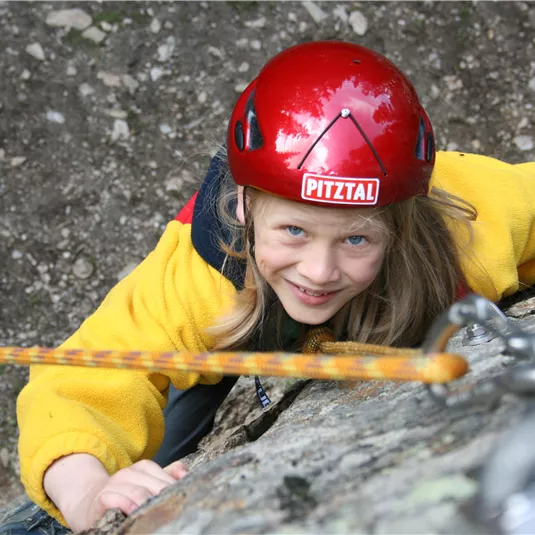 Ein junges Mädchen klettert an einem Felsen und trägt einen Helm. Sie lächelt und scheint viel Spaß zu haben.