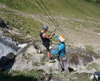 Two climbers are working on a rope beneath a waterfall. The surroundings are green and mountainous with flowing water.