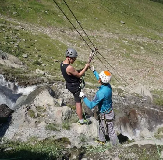 Two climbers are working on a rope beneath a waterfall. The surroundings are green and mountainous with flowing water.