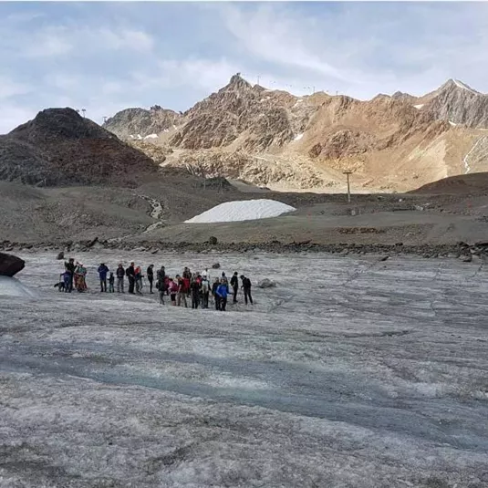 A group of people is standing on a glacier in a mountainous area. In the background, impressive mountains and a blue sky can be seen.