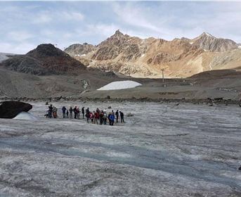 A group of people is standing on a glacier in the mountains. In the background, majestic mountain peaks and a clear sky can be seen.