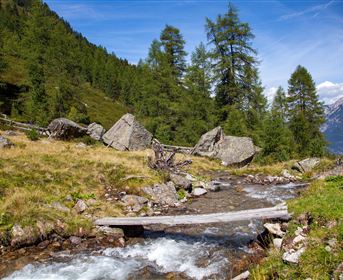 Eine Person steht auf einem Hügel und blickt auf die beeindruckende Berglandschaft. Im Hintergrund ist ein See und ein klarer Himmel mit einigen Wolken zu sehen.