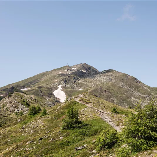 An impressive mountain landscape with gentle hills and snow-capped peaks. The sky is clear and blue, ideal for a hike in nature.
