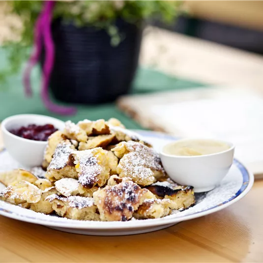 Ein Teller mit köstlichem Kaiserschmarrn, bestäubt mit Puderzucker. Dazu gibt es zwei kleine Schalen mit Marmelade und Vanillesauce.