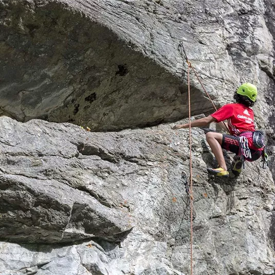A climber in a red shirt ascends a steep rock. He is wearing a helmet and climbing gear.