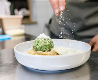 Ein Teller mit einer grünen, ballartigen Speise wird mit Käse bestreut. Im Hintergrund sind weitere Küchenutensilien sichtbar.