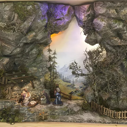 A detailed landscape depiction featuring a cave, trees, and a gentle sunset. In the foreground are figures reenacting a rural scene.