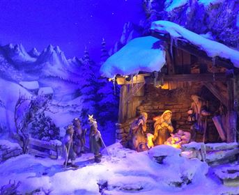 Eine winterliche Krippenszene mit schneebedeckten Bergen im Hintergrund. Die Figuren stehen um die Stallbeleuchtung, die eine warme Atmosphäre schafft.