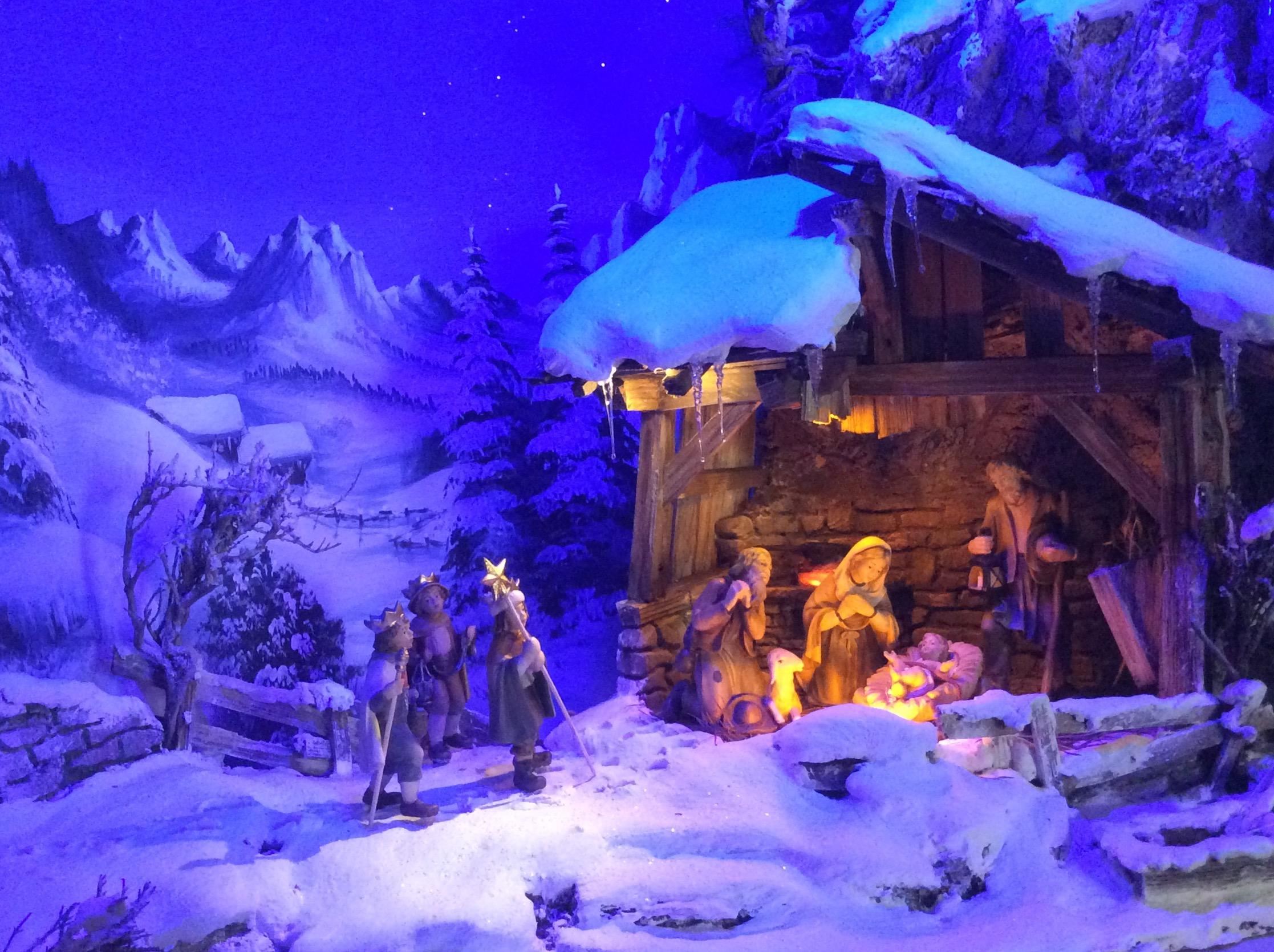 A wintry nativity scene with snow-covered mountains in the background. The figures stand around the stable lighting, which creates a warm atmosphere.