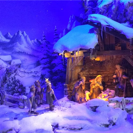A wintry nativity scene with snow-covered mountains in the background. The figures stand around the stable lighting, which creates a warm atmosphere.
