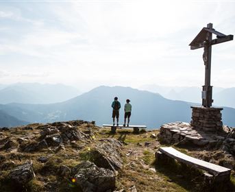 Zwei Menschen stehen auf einem Berggipfel neben einem Kreuz. Im Hintergrund erstreckt sich eine beeindruckende Berglandschaft mit einem klaren Himmel.