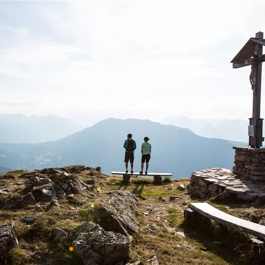 Zwei Menschen stehen auf einem Berggipfel neben einem Kreuz. Im Hintergrund erstreckt sich eine beeindruckende Berglandschaft mit einem klaren Himmel.