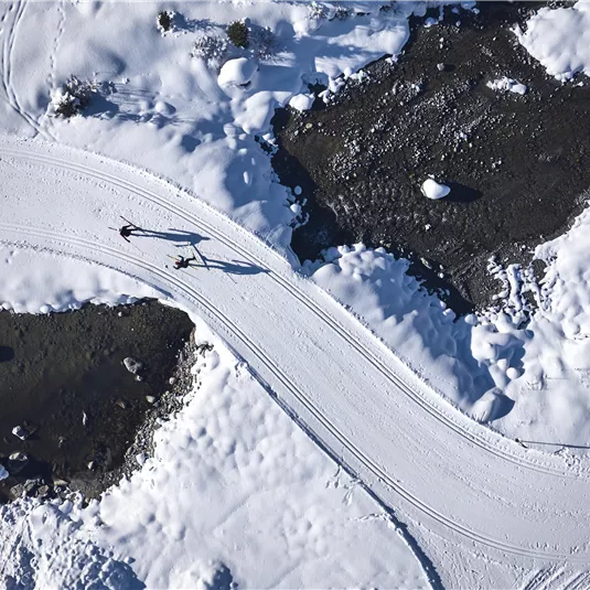 Eine kurvenreiche Schneestraße führt durch eine winterliche Landschaft. Im Hintergrund sind schneebedeckte Flächen und einige Felsen zu sehen.