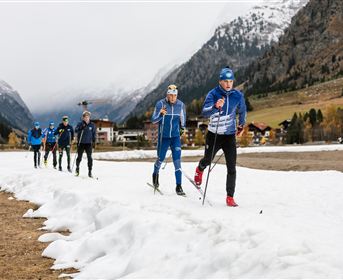 Eine Gruppe von Skilangläufern trainiert auf einer schneebedeckten Strecke in den Bergen. Im Hintergrund sind Berge und eine ländliche Landschaft zu sehen.