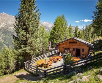 Eine charakteristische Almhütte umgeben von Bäumen. Es gibt Tische im Freien mit Sonnenschirmen und eine gut gepflegte Umgebung.