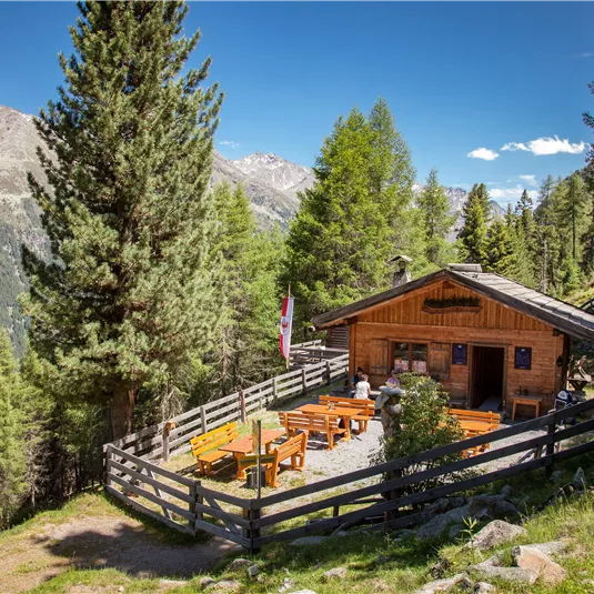 A characteristic alpine hut surrounded by trees. There are outdoor tables with umbrellas and a well-maintained environment.
